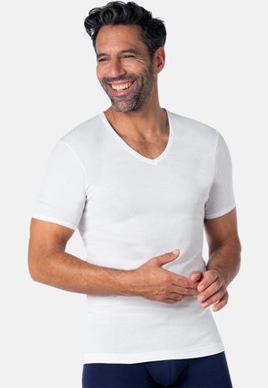 Homme souriant aux cheveux foncés et à la barbe, portant un t-shirt blanc ajusté à col en V et un pantalon bleu marine, debout devant un fond clair.