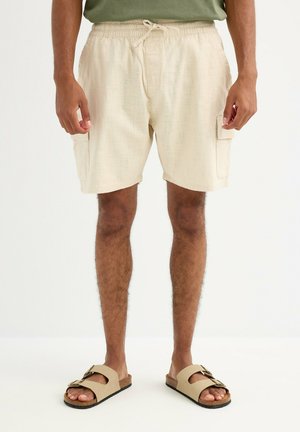 Hombre con pantalones cortos cargo beige con bolsillos laterales, camiseta verde y sandalias beige con hebilla, de pie sobre un suelo blanco.