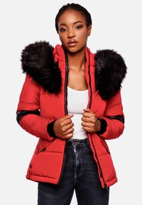 Chaqueta acolchada roja con cuello de piel sintética negra, cierre de cremallera y detalles en negro en las mangas. Lleva puesta una camiseta blanca y unos jeans oscuros.