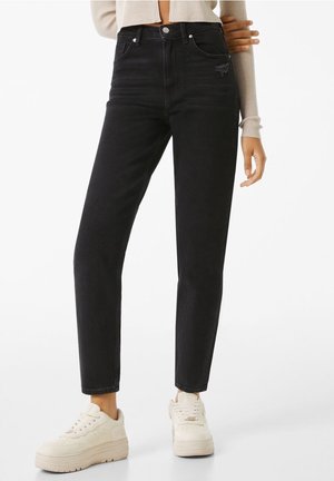 Kvinde iført sort high-waist jeans med let slid på lommen, beige cropped sweater og hvide platform sneakers mod hvid baggrund.
