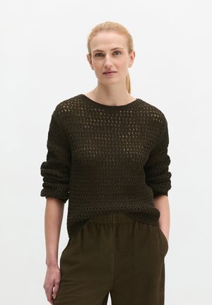 Femme aux cheveux blonds attachés en queue de cheval, portant un pull ajouré foncé et un pantalon sombre, une main dans la poche, debout devant un fond blanc.