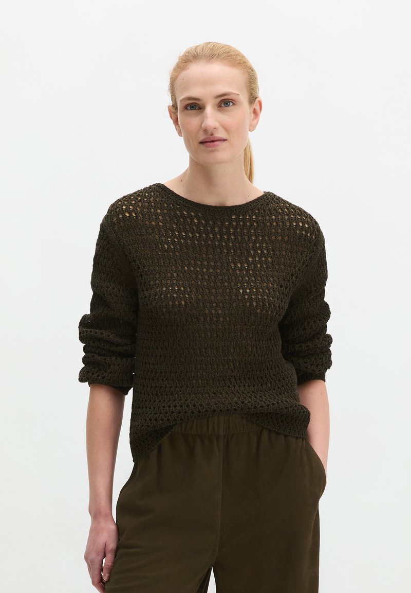Femme aux cheveux blonds attachés en queue de cheval, portant un pull ajouré foncé et un pantalon sombre, une main dans la poche, debout devant un fond blanc.
