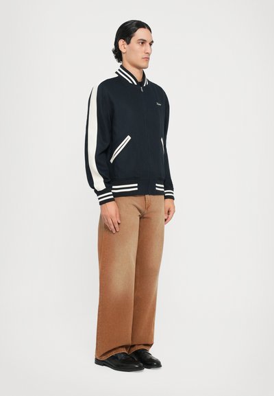 Veste aviateur marine avec des rayures blanches, poignets et taille côtelés. Associée à un pantalon large marron et des chaussures noires. Pas de logos visibles.