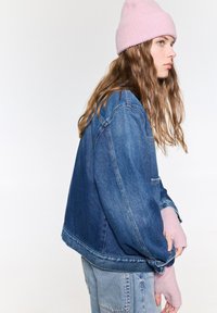 Giacca di denim blu medio, vestibilità oversize con tasca sul petto. Indossata con un berretto a coste rosa chiaro e guanti senza dita rosa.