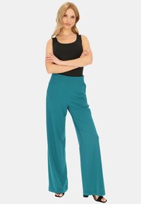 Turkusowe spodnie o szerokich nogawkach wykonane z gładkiego materiału z wygodnym pasem, w połączeniu z czarną bluzką bez rękawów, prezentowane na modelce.