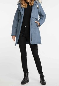 Parka azzurra chiara con cappuccio foderato in pelliccia, dotata di zip e bottoni, molteplici tasche e colletto leggermente rialzato. Pantaloni neri e stivali.