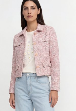 Frau trägt eine strukturierte pinke Jacke mit Knöpfen über einem weißen T-Shirt mit Blumenmuster und hellblauen Jeans, steht vor einem schlichten Hintergrund.