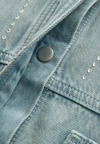 Jasnoniebieski jeansowy materiał z metalowym zatrzaskiem, precyzyjnym przeszyciem i małymi przezroczystymi ozdobnymi ćwiekami wzdłuż krawędzi materiału.