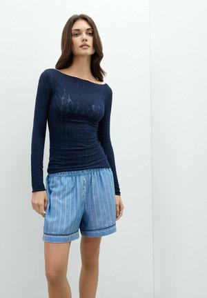 Mujer de pie que lleva una camiseta ajustada azul marino de manga larga y pantalones cortos sueltos azul claro con rayas verticales blancas y botones en la parte delantera.