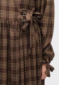 Hnedé kárované šaty s zavinovacím dizajnom, kovovým hardvérom s krúžkami a viazanými detailmi na rukávoch. Textúra je hladká a tkaná.
