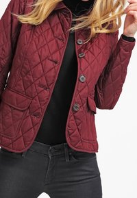 Bordeaux rode doorgestikte jas met knoopsluiting, voorzien van twee voorzakken en een getailleerde pasvorm, gedragen over een zwarte coltrui.