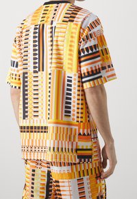 Lyhythihainen paita kirkkaan oranssina, keltaisina ja mustina kuvioina. Valmistettu kevyestä kankaasta, jossa on rohkeita geometrisiä muotoiluja ja kontrasteja.