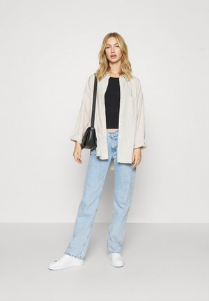 Camisa oversized às riscas beje, top crop preto, jeans retos azuis claros e sapatilhas brancas. Acessorizado com uma pequena mala preta.