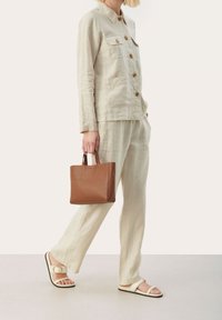 Veste en lin beige avec boutons en bois, pantalon assorti et sac fourre-tout en cuir marron. Sandales blanches avec une boucle décorative complètent la tenue.