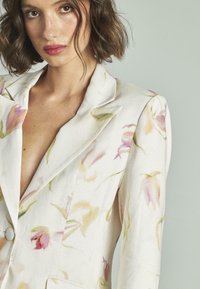 Blazer a medida de tela crema con un estampado floral en tonos rosa y verde. Detalles destacados incluyen un solapa y cierre de un solo botón.