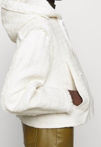 Biała, teksturowana bluza z kapturem, z sznurkiem, surowymi brzegami i bocznymi kieszeniami, noszona na brązowych skórzanych spodniach.