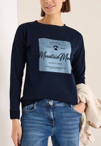 Tmavě modrá mikina s dlouhým rukávem, která má na sobě světle modrou grafiku s textem. Spárovaná se světle modrými džíny. Hladká textura a uvolněný střih.