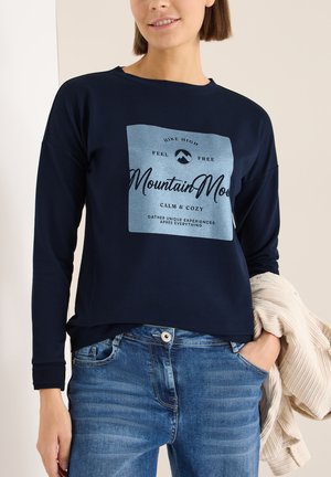 Tmavě modrá mikina s dlouhým rukávem, která má na sobě světle modrou grafiku s textem. Spárovaná se světle modrými džíny. Hladká textura a uvolněný střih.