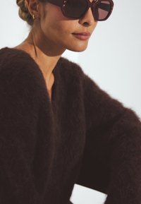 Maroon, fuzzy sweater with wide sleeves, paired with oversized, dark tinted sunglasses featuring decorative studs along the frame.