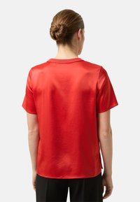 Rød top med korte ærmer lavet af glat, skinnende materiale. Enkel rund halsudskæring og afslappet pasform, med synlige syninger og en let buet kant.