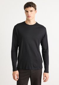 Ung mann iført en ensfarget svart langermet skjorte og mørke bukser, stående mot en hvit bakgrunn, vendt fremover med et nøytralt uttrykk.