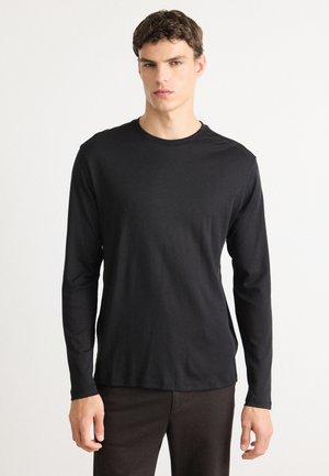 Un jeune homme portant une chemise noire unie à manches longues et un pantalon sombre, se tenant contre un fond blanc, faisant face à l'avant avec une expression neutre.