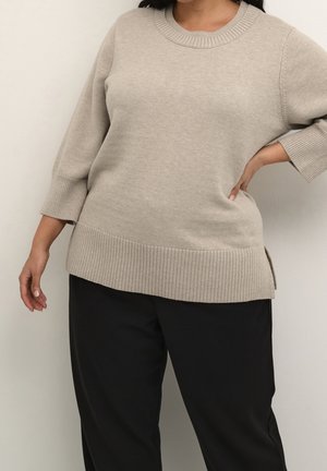 Vrouw draagt een beige gebreide trui met geribbelde manchetten en zoom, gecombineerd met een zwarte broek, met één hand op haar heup en de andere ontspannen langs haar lichaam.