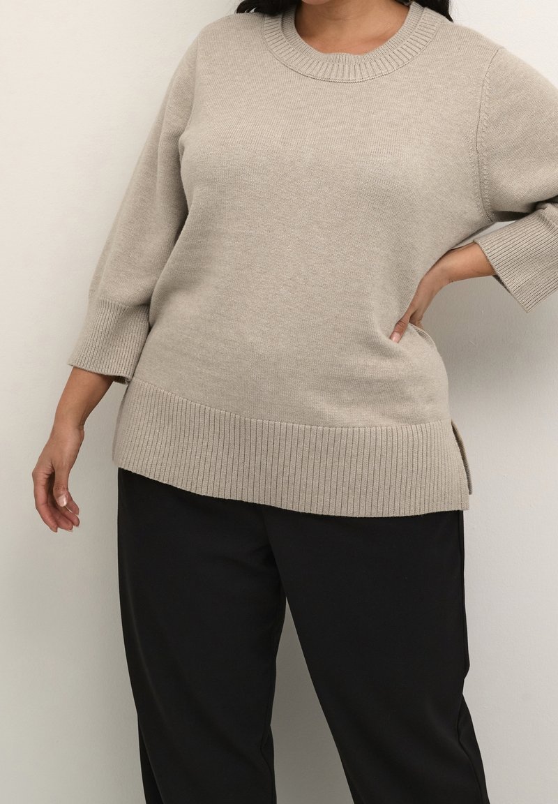 Femme portant un pull beige en maille avec poignets et ourlet côtelés, associé à un pantalon noir, une main sur la hanche et l'autre détendue le long du corps.