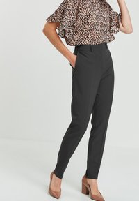 Pantalons tailleur noirs avec une coupe slim et des poches latérales, associés à un chemisier transparent à imprimé léopard avec des manches à volants. Chaussures à talons compensés marron.