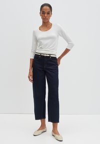 Weißes Langarmshirt, dunkelblaue weit geschnittene Jeans und cremefarbene Ballerinas mit strukturierter Musterung. Modell posiert vor einem schlichten Hintergrund.