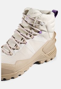 Beżowe buty trekkingowe z teksturowaną cholewką, wyściółką wokół kostki, fioletowymi akcentami i solidną podeszwą. Posiadają odblaskowe sznurówki oraz etykietę DryTechnology.