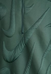 Πράσινο πουλόβερ με υφασμάτινη υφή, που διαθέτει μισό φερμουάρ στον γιακά και ανυψωμένο μοτίβο λογότυπου Nike σε διάφορες αποχρώσεις και σχήματα σε όλη την επιφάνεια.