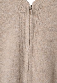 Cardigan en tricot beige avec fermeture éclair verticale, fabriqué en mélange de laine douce, présentant une texture subtile et de fines bordures en côtes le long des bords.
