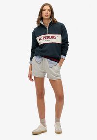 Granatowy sweatshirt z białym i czerwonym logo "SUPERDRY". Zestawiony z jasnoszarymi szortami. Model ma na sobie białe sneakersy i jasne skarpetki.