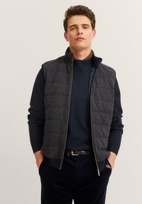 Gepolsterte Weste mit einem Reißverschluss vorne, aus dunkelgrauem strukturiertem Stoff, hoher Kragen und einem dezenten geometrischen Muster. Getragen über einem dunklen Pullover.