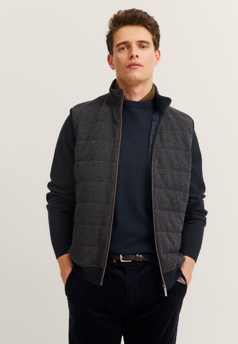 Gepolsterte Weste mit einem Reißverschluss vorne, aus dunkelgrauem strukturiertem Stoff, hoher Kragen und einem dezenten geometrischen Muster. Getragen über einem dunklen Pullover.