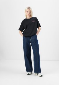 T-shirt en coton noir avec un col arrondi et des manches courtes, associé à un jean bleu à jambes larges. Détail notable du logo rose sur la poitrine.