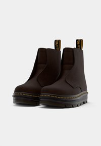 Bottines marron avec un matériau lisse, panneaux élastiques sur les côtés et coutures jaunes le long de la semelle noire. Elles disposent de languettes à l'arrière.