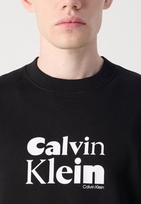 Black sweatshirt featuring a crew neck and ribbed detailing, with a prominent white “Calvin Klein” logo in varying font styles.