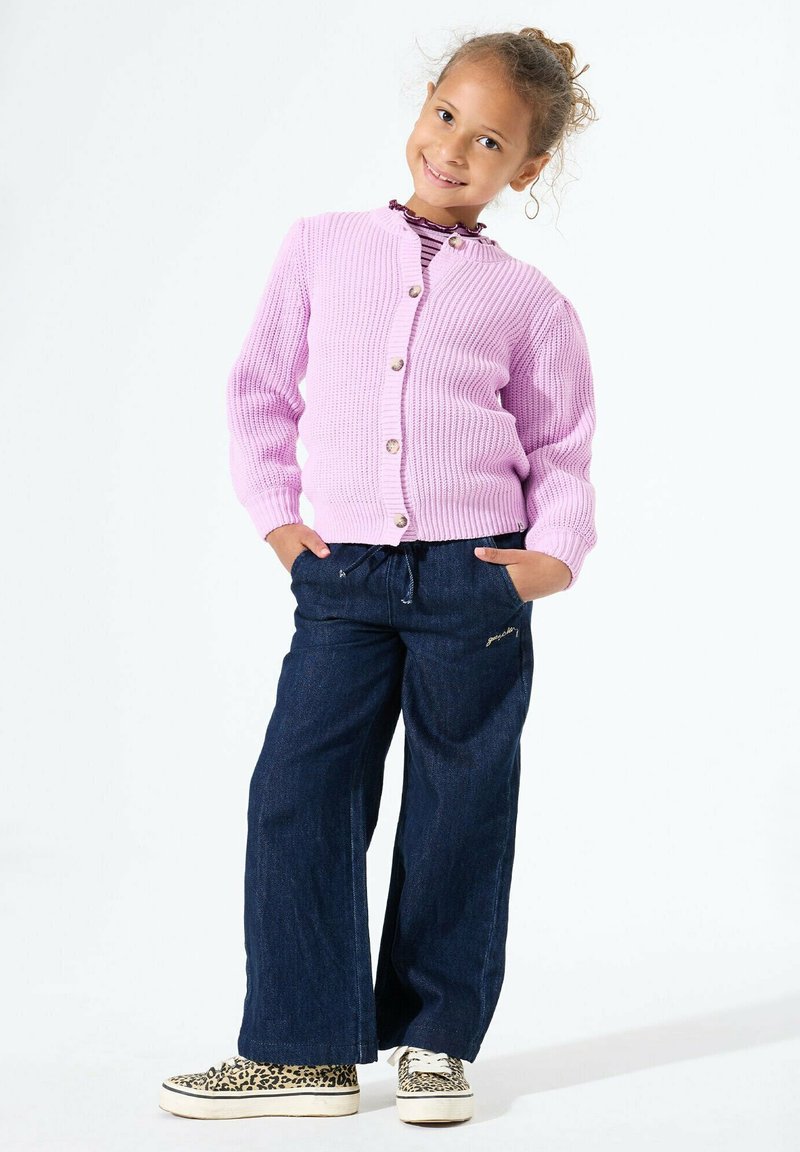 Lachend meisje met krullend haar draagt een roze cardigan met knopen, een gestreept shirt, wijde donkere jeans en sneakers met luipaardprint, met haar handen in haar zakken.