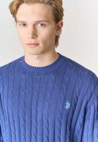 Maglione blu a coste con scollo a coste e piccolo emblema azzurro sul petto sinistro. Tessuto morbido e vestibilità comoda.