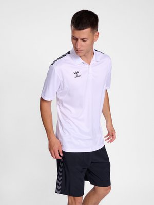 Jeune homme portant un polo blanc hummel et un short noir avec une bande chevron, debout et regardant vers le bas sur un fond uni.