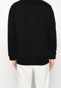 Felpa nera con scollo rotondo e maniche lunghe. Presenta polsini e orlo a costine. Texture liscia con design minimalista. Indossata sopra pantaloni bianchi.