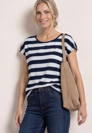 Smiling woman with blonde hair in a navy and white striped shirt, dark blue jeans, holding a tan suede shoulder bag against a light wall.