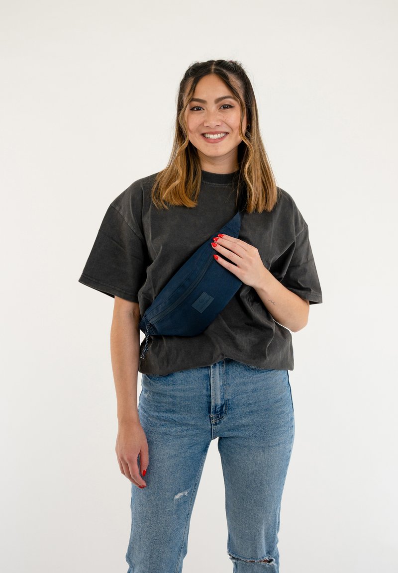 Navyblaue Gürteltasche mit glatter Oberfläche und verstellbarem Gürtel, über einem schwarzen T-Shirt und hellblauen Jeans quer getragen.