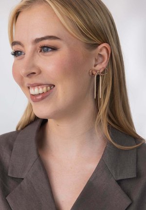 Ohrringe aus Roségold mit einer Schleifendesign und zwei langen, schlanken Metallsträngen. Das Modell trägt ein graues Oberteil mit Kragen und strukturierter Oberfläche.