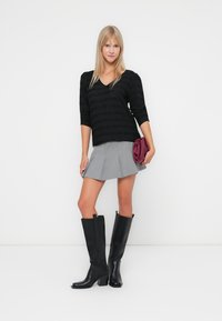 Zwarte, gestructureerde top, grijze geplooide rok en kniehoge zwartleren laarzen. Houdt een bordeauxrode clutch vast. Eenvoudig, modern ontwerp en contrasterende kleuren.