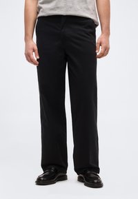 Άνδρας που φοράει μαύρο παντελόνι ίσιας γραμμής, μαύρα δερμάτινα παπούτσια και γκρι κοντομάνικο πουκάμισο, στέκεται μπροστά από απλό λευκό φόντο.