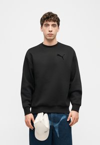 Νεαρός άνδρας που φοράει μαύρο φούτερ Puma και μπλε τζιν, κρατώντας ένα λευκό καπέλο Puma μπροστά σε απλό λευκό φόντο.