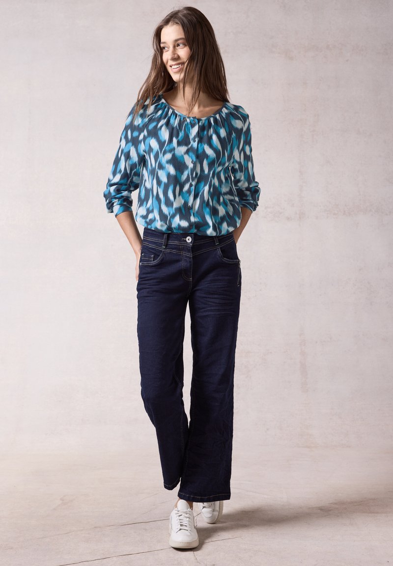 Blusa blu e bianca a fantasia con scollatura a barca, abbinata a jeans blu scuro e sneakers bianche. La modella sta in piedi con le mani nelle tasche.